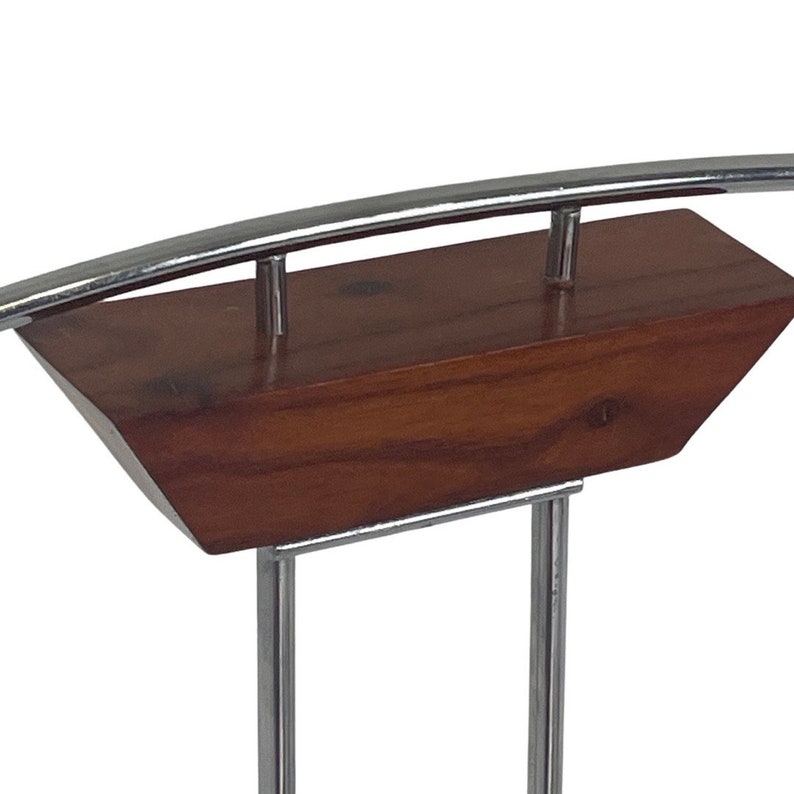 May include: A chrome and wood magazine rack with a curved top bar and two vertical supports. The rack has a rectangular wooden base with a beveled edge.