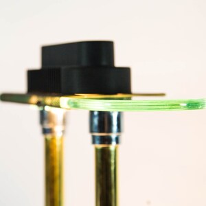 May include: Close-up of a modern, abstract lamp design. The lamp features a black rectangular top, a curved, light green glass element, and two gold-colored metal supports. The design is minimalist and contemporary, with a focus on clean lines and contrasting colors.