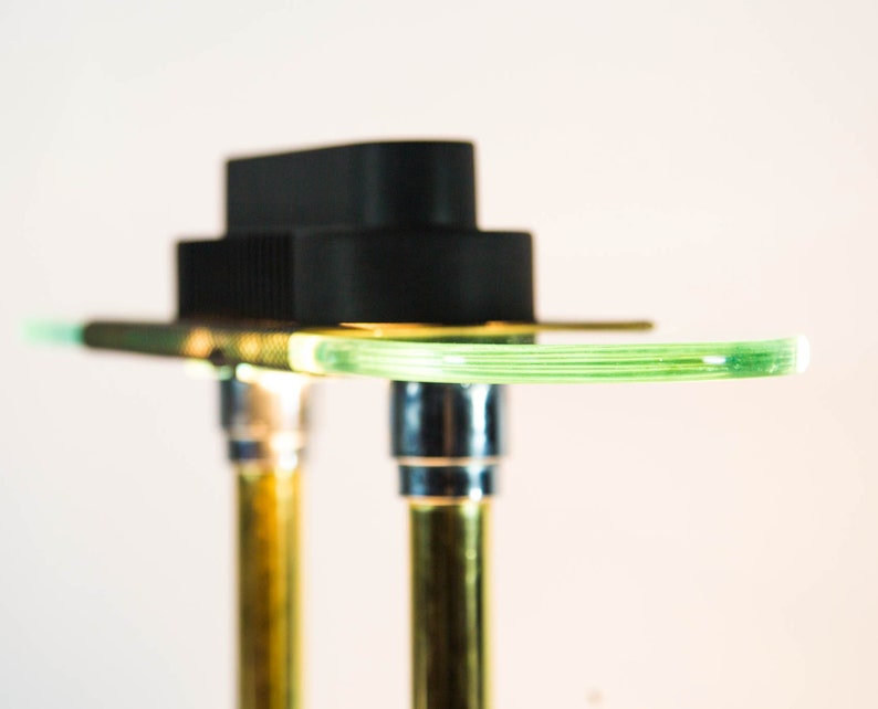 May include: Close-up of a modern, abstract lamp design. The lamp features a black rectangular top, a curved, light green glass element, and two gold-colored metal supports. The design is minimalist and contemporary, with a focus on clean lines and contrasting colors.