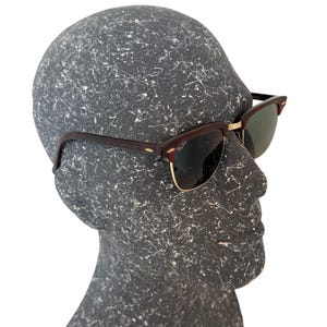 May include: Sunglasses with dark brown frames and gold-colored accents. The lenses are a dark green color. The sunglasses are displayed on a gray mannequin head.