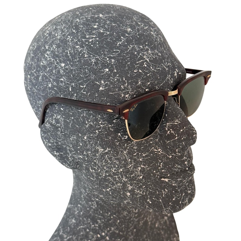 May include: Sunglasses with dark brown frames and gold-colored accents. The lenses are a dark green color. The sunglasses are displayed on a gray mannequin head.