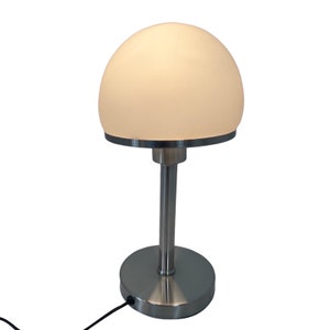 May include: A modern table lamp with a frosted, dome-shaped shade. The lamp has a brushed metal base, a slender stem, and a circular base. The shade emits a soft, warm light. The lamp is connected to a power source via a black cord.