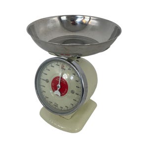 May include: A vintage white kitchen scale with a chrome bowl and a silver face. The scale has a red dial with black markings and the text "CAPACITY 1.2 KG" and "SENSITIVITY 10G".