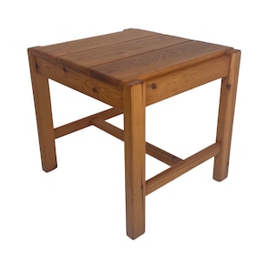 Op de afbeelding: Een kleine, vierkante houten tafel met een eenvoudig ontwerp. De tafel is gemaakt van lichtbruin hout en heeft een natuurlijke afwerking. Het heeft een enkele plank onder het tafelblad.