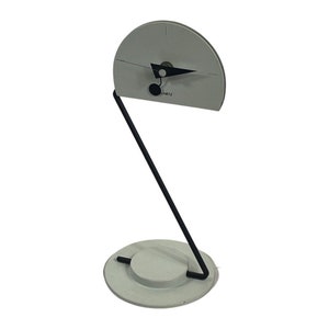 Puede incluir: Un reloj blanco con agujas negras y una etiqueta negra "QUARTZ" en un soporte de metal negro. La esfera del reloj es un círculo con un puntero triangular negro.