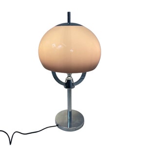 May include: A vintage table lamp with a chrome base and stem, supporting a round, peach-coloured glass shade. The lamp features a black accent at the top and a curved chrome support. A black power cord extends from the base.