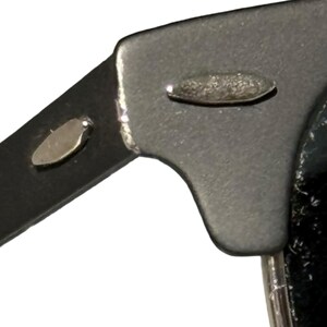 May include: Detail of a black sunglasses frame with silver metal rivets. The frame has a matte finish and features two small, oval-shaped silver rivets on the arm and hinge area. The design is simple and modern.