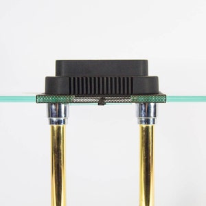 May include: A modern side table with a clear glass top and a black rectangular object on top. The table has two gold-colored legs with chrome accents. The table is approximately 24 inches wide.