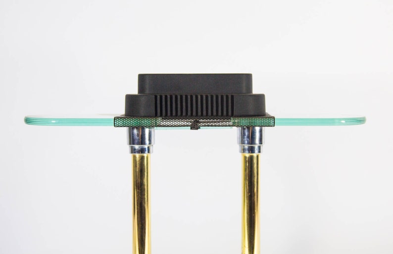 May include: A modern side table with a clear glass top and a black rectangular object on top. The table has two gold-colored legs with chrome accents. The table is approximately 24 inches wide.