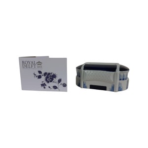 May include: A small, white ceramic box with blue and white details. The box is shaped like a building and has a blue roof. The box is labelled "Royal Delft 1653" and has a small crown above the word "Royal".