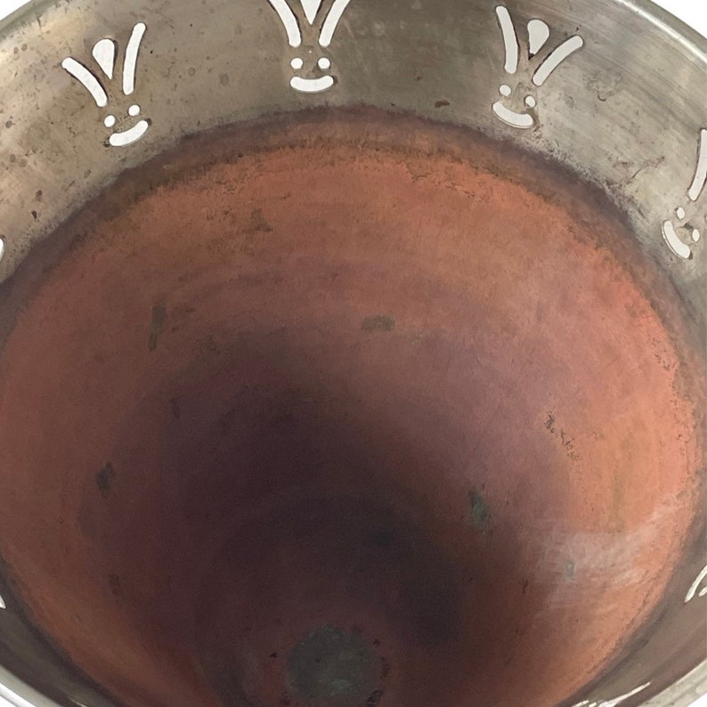 Pu&ograve; includere: Una ciotola d'argento con interno in rame. La ciotola ha un design decorativo attorno al bordo con corone stilizzate bianche.