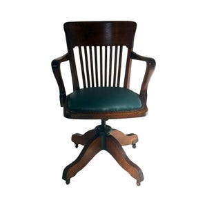 May include: An antique wooden office chair with a dark brown finish. The chair features a green leather seat, a slatted back, and curved armrests. It has a five-star base with small wheels for mobility, and a swivel mechanism.