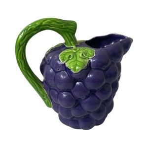 May include: A purple ceramic pitcher shaped like a bunch of grapes with green leaves. The pitcher has a handle and a spout.
