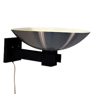 Op de afbeelding: Een zwarte metalen wandlamp met een zilveren, komvormige kap. De lamp heeft een eenvoudig, modern ontwerp en is perfect om elke kamer een vleugje stijl te geven.