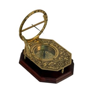 May include: A brass sundial compass with an etched floral design on a dark wood base. The compass features a circular dial with a needle and a hinged gnomon. The sundial has Roman numerals and a curved arm for measuring time.