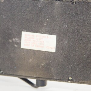 May include: A close-up of a black rectangular object with a white label. The label reads "ROXFORD HOLLAND B.V. HALOGEN LIGHT" and includes model and voltage information. The object has a textured surface and a small, curved metal piece.