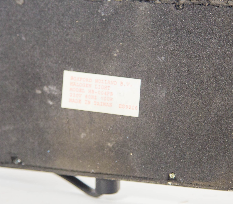 May include: A close-up of a black rectangular object with a white label. The label reads "ROXFORD HOLLAND B.V. HALOGEN LIGHT" and includes model and voltage information. The object has a textured surface and a small, curved metal piece.