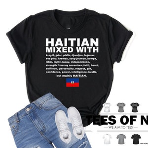 Puede incluir: Camiseta negra con texto blanco que dice "Haitian Mixed With" y enumera varias palabras y frases haitianas. El texto tambi&eacute;n incluye la bandera haitiana. La camiseta est&aacute; sobre un fondo blanco con un par de jeans y zapatillas blancas.
