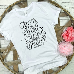 Puede incluir: Camiseta blanca con texto negro que dice "She is far more precious than jewels Proverbs 31:10".