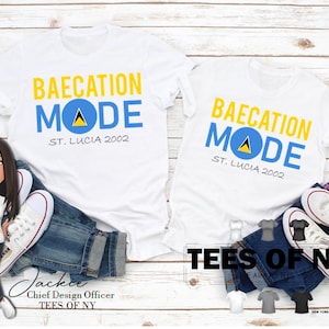 May include: Two white t-shirts with the text "Bae-cation Mode" and the flag of Saint Lucia. The text "St. Lucia 2002" is below the flag. The shirts are on a white wood background.