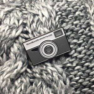 May include: A black and white enamel pin of a vintage camera with the text "СМЕНА-РАПИД" on the front.