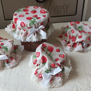 Peut inclure: Quatre bocaux en verre surmontés de couvercles en tissu à motif de fraises rouges sur fond crème. Chaque housse est agrémentée d'une bordure en dentelle blanche et d'un petit nœud en ruban blanc fixé à l'avant sur le couvercle du bocal.