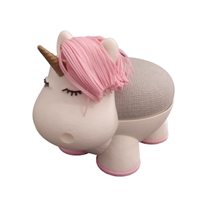 May include: A white unicorn-shaped speaker holder with pink accents. The holder features a gold horn, pink mane, and closed eyes. The speaker is gray and fits into the unicorn's back. The base and hooves are outlined in pink.