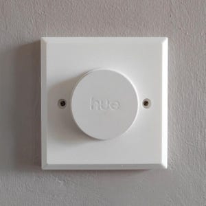 May include: A white square wall-mounted smart light dimmer switch featuring a circular centre dial with the word hue embossed in the middle. The device is secured to the wall with two visible screws on the left and right sides of the dial.