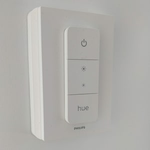 May include: White Philips Hue smart light switch with a power button, two brightness controls, and the word "hue" printed on the face. The switch is mounted on a white wall.