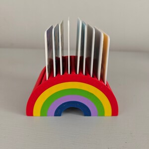 Puede incluir: Un tarjetero en forma de arco&iacute;ris, rojo en la parte superior y con un dise&ntilde;o de arco&iacute;ris en amarillo, verde, morado y azul. El soporte contiene varias tarjetas blancas. El soporte es de pl&aacute;stico.
