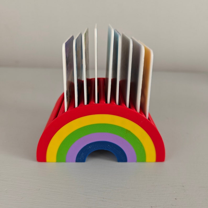 Puede incluir: Un tarjetero en forma de arco&iacute;ris, rojo en la parte superior y con un dise&ntilde;o de arco&iacute;ris en amarillo, verde, morado y azul. El soporte contiene varias tarjetas blancas. El soporte es de pl&aacute;stico.