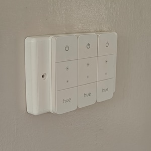 May include: A white smart home control panel fixed to a light grey wall. The panel features nine square buttons with power, snowflake, and "hue" symbols. It is designed for controlling smart home devices.
