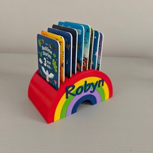 Puede incluir: Un sujetalibros en forma de arco&iacute;ris rojo, amarillo, verde, morado y azul, con el nombre "Robyn" en azul. Contiene varios libros infantiles con cubiertas coloridas. El sujetalibros est&aacute; dise&ntilde;ado para colocarse sobre una superficie plana.
