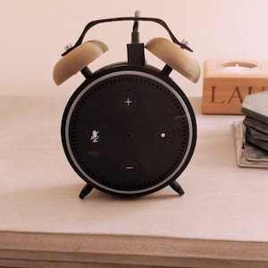 Op de afbeelding: Een zwarte Amazon Echo Dot slimme speaker met een 3D-geprinte wekkerstandaard. De standaard heeft twee beige, klokvormige toppen en een zwarte basis.