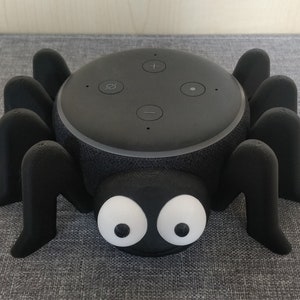 May include: A black Amazon Echo Dot smart speaker in a 3D printed black spider stand with white eyes.