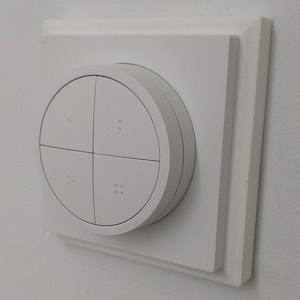 May include: A white, square-shaped wall switch with a round, white button in the centre. The button has four sections, each with a small, raised dot.