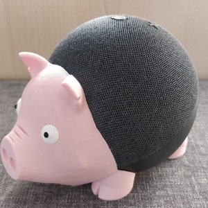 Peut inclure: Une housse de haut-parleur en forme de cochon rose pour un haut-parleur intelligent noir. La housse de haut-parleur a une surface en tissu texturé et des yeux blancs.