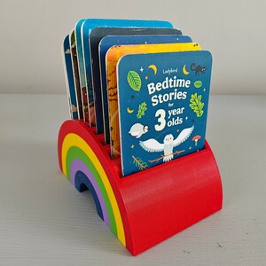 Puede incluir: Un sujetalibros rojo en forma de arco&iacute;ris con una colecci&oacute;n de libros infantiles. Los libros, titulados "Bedtime Stories for 3 year olds", presentan ilustraciones de animales y escenas nocturnas. El soporte es de madera.