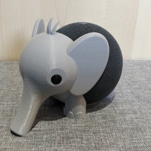 Peut inclure: Un support d'éléphant imprimé en 3D de couleur grise pour un haut-parleur intelligent noir. L'éléphant a une longue trompe et de grandes oreilles.