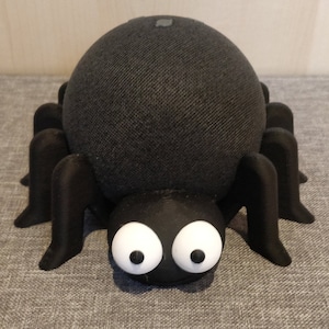 Peut inclure: Un haut-parleur noir recouvert de tissu avec un support en forme d'araignée. Le support a huit pattes et de grands yeux blancs.