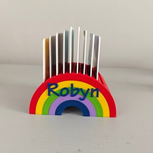 Puede incluir: Un tarjetero en forma de arco&iacute;ris, rojo, amarillo, verde, morado y azul, con el nombre "Robyn" en azul. Contiene varias tarjetas. El dise&ntilde;o es brillante y alegre.