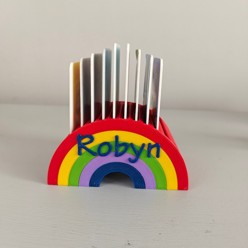 Puede incluir: Un tarjetero en forma de arco&iacute;ris, rojo, amarillo, verde, morado y azul, con el nombre "Robyn" en azul. Contiene varias tarjetas. El dise&ntilde;o es brillante y alegre.