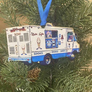 May include: Wooden ornament shaped like an ice cream truck, featuring the text "Mister Softee" and "Sundaes, Shakes, Cones." The truck is white with blue accents and a blue ribbon for hanging. The ornament is set against a backdrop of green pine needles and a pine cone.