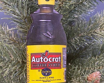 May include: A Christmas tree ornament shaped like an Autocrat Coffee Syrup bottle. The ornament has a yellow label with red text and a red bird logo. The bottle is dark brown with a yellow cap and a yellow ribbon for hanging.
