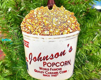 May include: A popcorn-shaped Christmas ornament. The ornament is a white bucket filled with caramel popcorn. The bucket has the words "Johnson's Popcorn" printed on it, along with "World Famous Quality Caramel Corn Since 1940". A brown ribbon is attached.