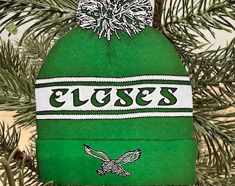 May include: Green Christmas ornament shaped like a beanie hat. The hat has a white band with the word "EAGLES" in green letters. A green and white pom-pom tops the hat, and a green ribbon is attached for hanging. An eagle logo is on the front.