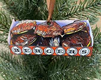 May include: A Christmas bauble shaped like a box of Bergers Cookies. The box is red and white with a blue background. The cookies are brown and have a realistic texture. The bauble has a brown ribbon for hanging.