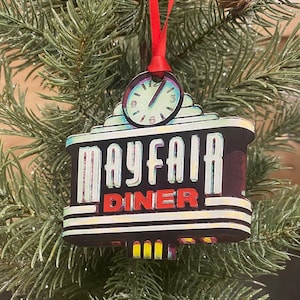 May include: A Mayfair Diner Christmas ornament with a clock design at the top. The ornament is rectangular with the words "MAYFAIR DINER" in white and red lettering. It hangs from a red ribbon on a Christmas tree.