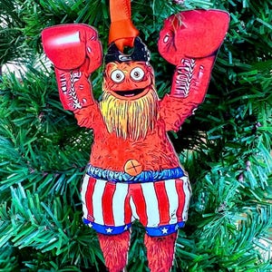 May include: A festive Christmas ornament featuring a cartoon character in boxing attire. The ornament is red with a yellow beard, wearing boxing gloves and patriotic shorts with stars and stripes. It hangs from an orange ribbon.