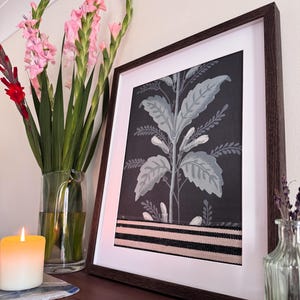 May include: A framed botanical print featuring a dark background with a stylized plant design in shades of gray and white. The print is accented with a black and tan striped band. A lit candle and pink flowers in a vase are in the foreground.
